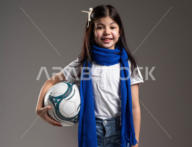 النظر إلى الكاميرا بإيماءات الفرح و السرور, بورتريه لفتاة عربية سعودية خليجية ترتدي الوشاح الأزرق و تحمل بيدها كرة القدم, تشجيع الأندية الرياضية السعودية, الشغف في ممارسة الرياضة المفضلة منذ الصغر, أنشطة ترفيهية للأطفال, خلفية رمادية.