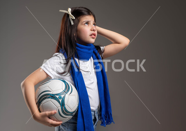 Looking up with gestures of confusion and sadness, portrait of a Saudi Gulf Arab girl wearing a blue scarf and holding a soccer ball in her hand, expressions of concern and bewilderment, recreational sports activities for children, passion for soccer since childhood, gray background.