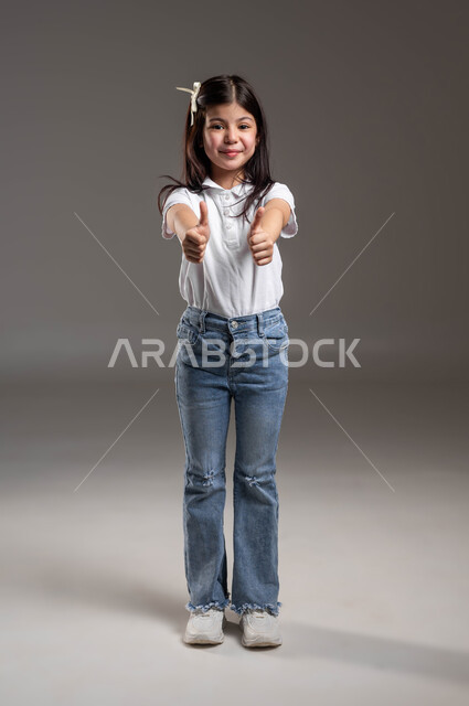 الدلالة على الإمتياز والتميز ،الموافقة على أمر معين، بورتريه لفتاة عربية خليجية سعودية مبتسمة ترتدي الزي الكاجوال ترفع اصابع الإبهام للاعلى بتعابير علامة الجودة، النظر إلى الكاميرا بايماءات الإعجاب والشعور بالسعادة، صورة الجسم بالطول الكامل، خلفية رمادية