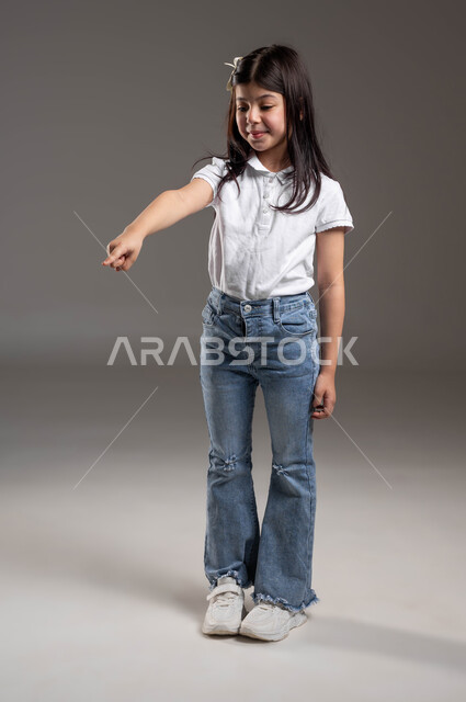 ايماءات الفرح لوجود شيء معين، حركات تدل على هذا المكان (هنا)، بورتريه لفتاة عربية خليجية سعودية يرتدي الزي الكاجوال تنظر إلى الأسفل وتشير باصبع السبابة الى شيء ما، تعابير تدل على السعادة والسرور، صورة الجسم بالطول الكامل، خلفية رمادية