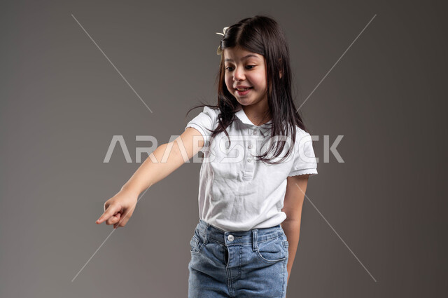 حركات تدل على هذا المكان (هنا)، ايماءات الفرح لوجود شيء معين، بورتريه لفتاة عربية خليجية سعودية ترتدي الزي الكاجوال تنظر إلى الأسفل وتشير باصبع السبابة الى شيء ما، تعابير تدل على السعادة والسرور، خلفية رمادية