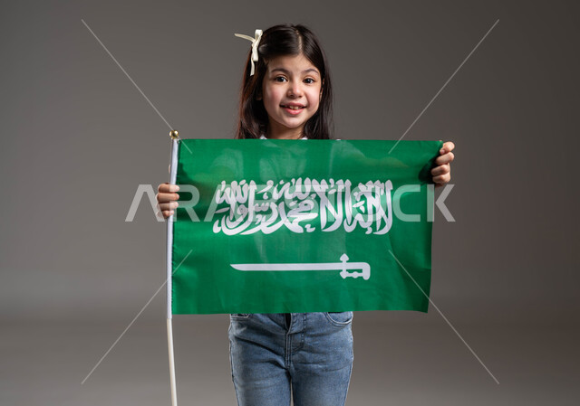 Looking at the camera with gestures of happiness and pleasure, commemorating Saudi National Day, September 23, Flag Day, March 11, portrait of a Saudi Gulf Arab girl wearing a casual outfit, holding the Kingdom’s flag in her hand, with expressions of pride and pride in national identity, gray background.