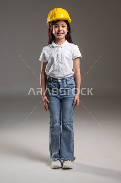 مفهوم الهندسة المدنية والمعمارية، بورتريه لبنت عربية خليجية سعودية مبتسمة ترتدي الزي الكاجوال و خوذة الوقاية الخاصة بالعمل تنظر إلى الكاميرا بايماءات السعادة والسرور، الشغف بمهنة الهندسة منذ الطفولة، مهن ووظائف مستقبلية، الجسم بالطول الكامل ,خلفية رمادية
