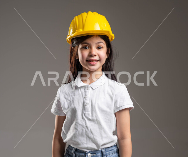 الشغف بمهنة الهندسة منذ الطفولة، مفهوم الهندسة المدنية والمعمارية، بورتريه لبنت عربية خليجية سعودية مبتسمة ترتدي الزي الكاجوال و خوذة الوقاية الخاصة بالعمل تنظر إلى الكاميرا بايماءات السعادة والسرور، مهن ووظائف مستقبلية، خلفية رمادية
