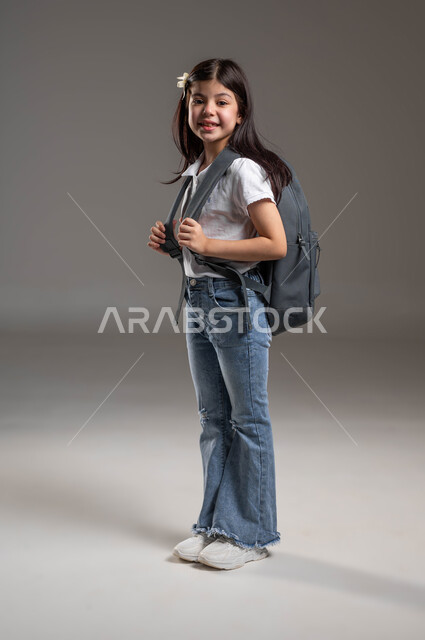 الاستعداد لبدء العام الدراسي الجديد، النظر الى الكاميرا بايماءات الفرح والسرور، مفهوم التعليم في السعودية، بورتريه لطالبة عربية خليجية سعودية مبتسمة ترتدي الزي الكاجوال وتحمل حقيبة الظهر المدرسية، المثابرة لتحقيق التفوق والنجاح، الطول الكامل للجسم، خلفية رمادية