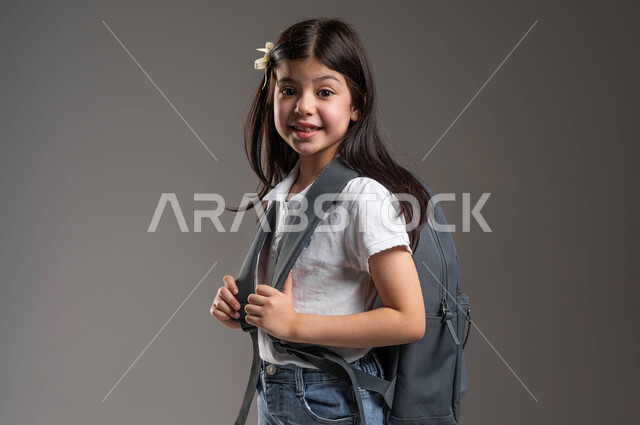 النظر الى الكاميرا بايماءات الفرح والسرور، المثابرة لتحقيق التفوق والنجاح،  مفهوم التعليم في السعودية، بورتريه لطالبة عربية خليجية سعودية مبتسمة ترتدي الزي الكاجوال وتحمل حقيبة الظهر المدرسية، الاستعداد لبدء العام الدراسي الجديد، خلفية رمادية