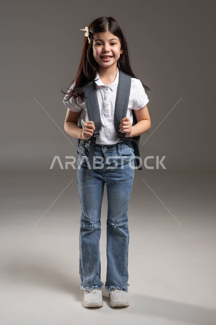 المثابرة لتحقيق التفوق والنجاح، التجهز لبدء العام الدراسي الجديد، النظر الى الكاميرا بايماءات الفرح والسرور، مفهوم التعليم في السعودية، بورتريه لطالبة عربية خليجية سعودية مبتسمة ترتدي الزي الكاجوال وتحمل حقيبة الظهر المدرسية، صورة الجسم بالطول الكامل, خلفية رمادية
