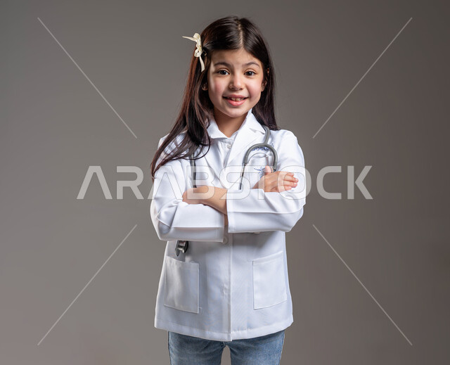 الطموح في تحقيق الآمال والأمنيات، الشغف بالحصول على مهنة الطب منذ الصغر، الوقوف باستقامة و النظر الى الكاميرا بإيماءات أيدي متقاطعة، بورتريه لفتاة عربية خليجية سعودية مبتسمة ترتدي المعطف الأبيض وسماعة الطبيب، خلفية رمادية