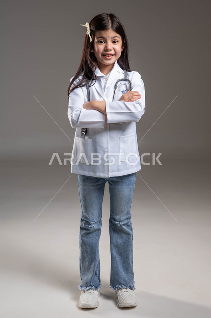 الوقوف باستقامة و النظر الى الكاميرا بإيماءات أيدي متقاطعة، الطموح في تحقيق الآمال والأمنيات، الشغف بالحصول على مهنة الطب منذ الصغر، بورتريه لفتاة عربية خليجية سعودية مبتسمة ترتدي المعطف الأبيض وسماعة الطبيب، صورة الجسم بالطول الكامل، خلفية رمادية