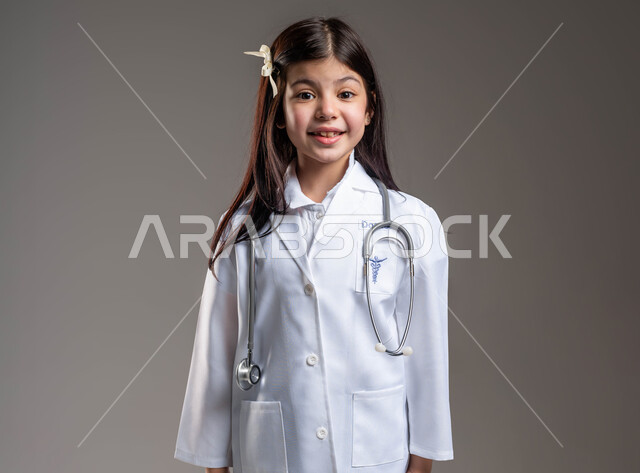 الشغف بالحصول على مهنة الطب منذ الصغر، بورتريه لفتاة عربية خليجية سعودية مبتسمة ترتدي المعطف الأبيض وسماعة الطبيب وتنظر إلى الكاميرا بإيماءات الفرح والسعادة، تحقيق الآمال والأمنيات من خلال الإجتهاد الدراسي، خلفية رمادية