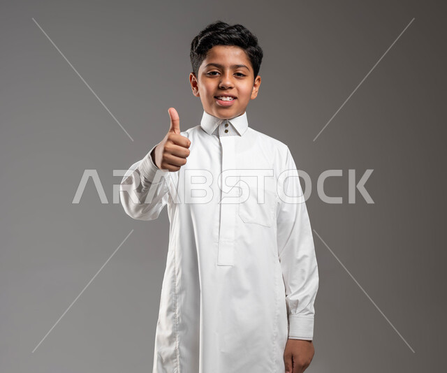 الإمتياز والتميز وإتقان العمل، رفع أصبع الإبهام دليل على علامة الجودة، الموافقة على موضوع ما، بورتريه لولد عربي خليجي سعودي مبتسم و يرتدي الزي التقليدي، ايماءات الإعجاب والرضا والشعور بالسعادة، خلفية رمادية