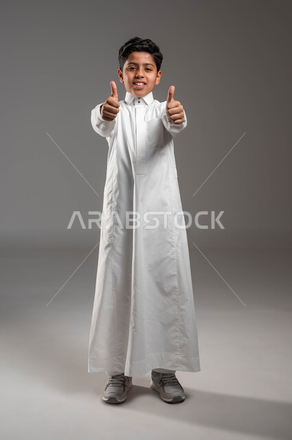 النظر إلى الكاميرا بايماءات الإعجاب والرضا والشعور بالسعادة، الموافقة على موضوع ما، بورتريه لفتى عربي خليجي سعودي مبتسم يرتدي الزي التقليدي يرفع اصابع الإبهام بتعابير علامة الجودة، الدلالة على الإمتياز والتميز، صورة الجسم بالطول الكامل، خلفية رمادية