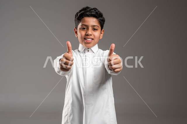 الموافقة على أمر معين، بورتريه مقرب لفتى عربي خليجي سعودي مبتسم يرتدي الزي التقليدي يرفع اصابع الإبهام بتعابير علامة الجودة، النظر إلى الكاميرا بايماءات الإعجاب والرضا والشعور بالسعادة، الدلالة على الإمتياز والتميز، خلفية رمادية