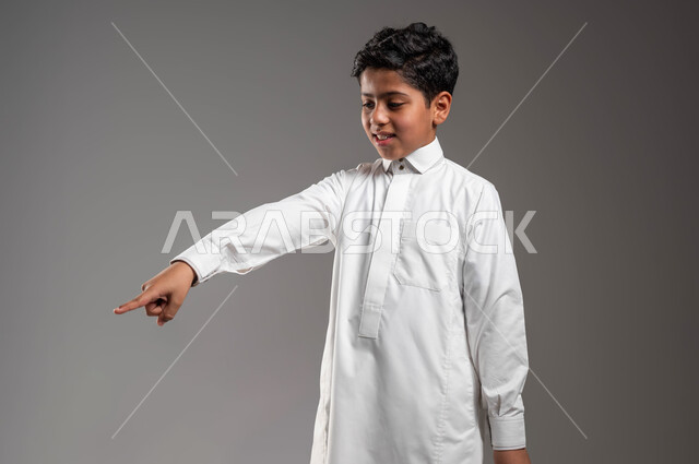 تعابير تدل على السعادة والسرور، ايماءات الفرح لوجود شيء معين، حركات تدل على هذا المكان (هنا)، بورتريه لفتى عربي خليجي سعودي يرتدي الثوب التقليدي ينظر إلى الأسفل ويشير باصبع السبابة الى شيء ما، خلفية رمادية