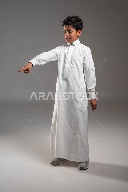 Gestures of joy for the presence of a certain thing, movements indicating this place (here), portrait of a Saudi Gulf Arab boy wearing a traditional dress, looking down and pointing with his index finger at something, expressions indicating happiness and pleasure, full-length body image, gray background