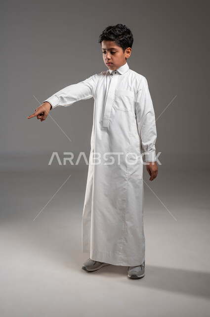 ايماءات تحديد موقع معين، حركات تدل على هذا المكان (هنا)، بورتريه لفتى عربي خليجي سعودي يرتدي الثوب التقليدي ينظر إلى الأسفل ويشير باصبع السبابة الى شيء ما، تعابير تدل على عدم الإعجاب والعبوس، صورة الجسم بالطول الكامل، خلفية رمادية