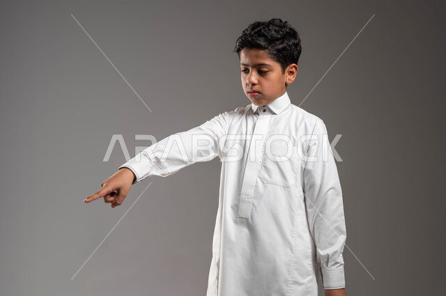 حركات تدل على هذا المكان (هنا)، بورتريه لفتى عربي خليجي سعودي يرتدي الثوب التقليدي ينظر إلى الأسفل ويشير باصبع السبابة الى شيء ما، تعابير تدل على عدم الإعجاب والعبوس، ايماءات تحديد موقع معين، خلفية رمادية