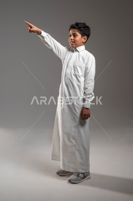 حركات تدل على تحديد موقع معين، مفهوم رؤية الأشياء البعيدة، تعابير وايماءات تدل على السرور، بورتريه لفتى عربي خليجي سعودي يرتدي الزي التقليدي يرفع يده الى الأعلى ويشير الى مكان ما بتعابير السرور، ايماءات الفرح لمشاهدة شيء، صورة الجسم بالطول الكامل، خلفية رمادية