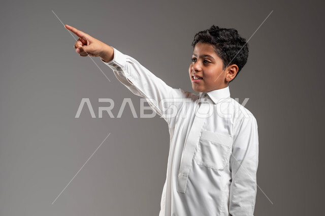 مفهوم رؤية الأشياء البعيدة، تعابير وايماءات تدل على السرور، بورتريه مقرب لفتى عربي خليجي سعودي يرتدي الزي التقليدي يرفع يده الى الأعلى ويشير الى مكان ما بتعابير السرور، حركات تدل على تحديد موقع معين، ايماءات الفرح لمشاهدة شيء، خلفية رمادية