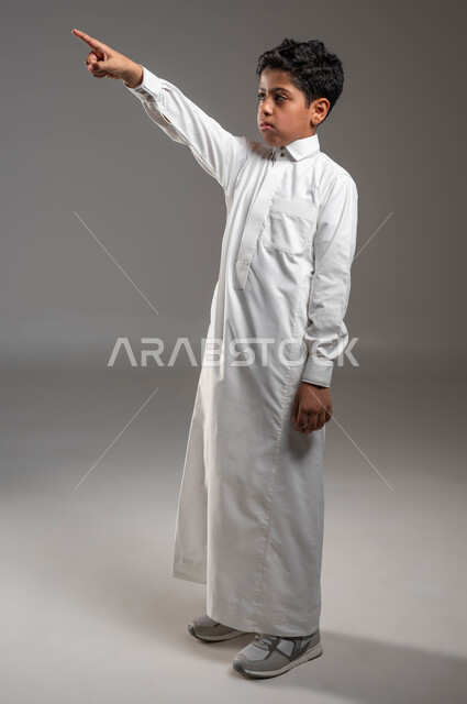 لفت الإنتباه إلى شيء معين، بورتريه لفتى عربي خليجي سعودي يرتدي الزي التقليدي يقوم بالنظر والإشارة الى شيء ما، توجيه الأصبع السبابة إلى الأعلى، ايماءات العبوس وعدم الإعجاب، رؤية الأشياء البعيدة، الجسم بالطول الكامل، خلفية رمادية