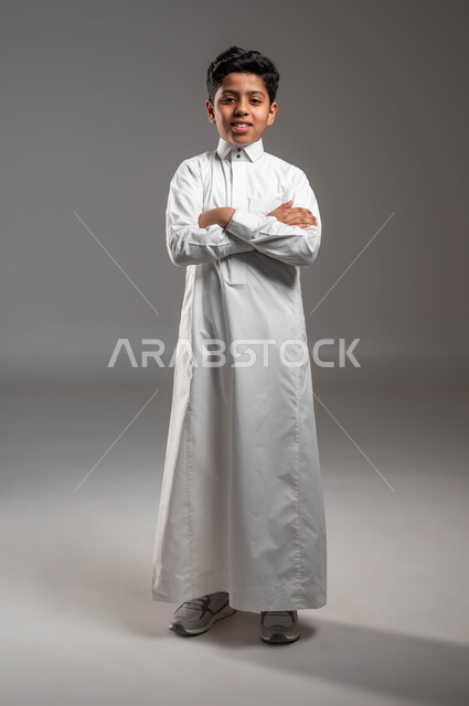 مفهوم الاهتمام بالمظهر الخارجي, الوقوف باستقامة بإيماءات أيدي متقاطعة، بورتريه لولد عربي خليجي سعودي مبتسم يرتدي الثوب التقليدي ينظر الى الكاميرا بتعابير الفرح والسرور، صورة الجسم بالطول الكامل، خلفية رمادية