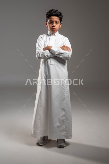 Gestures of boredom while waiting, looking at the camera with expressions of despair and dissatisfaction, portrait of a Saudi Gulf Arab boy wearing traditional clothing, standing straight with crossed hand movements, full length body portrait, gray background.
