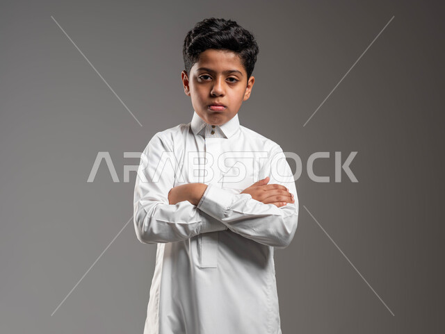 النظر الى الكاميرا بتعابير اليأس وعدم الرضا، بورتريه لولد عربي خليجي سعودي يرتدي الزي التقليدي يقف باستقامة بحركات ايدي متقاطعة، ايماءات الملل أثناء الإنتظار, خلفية رمادية