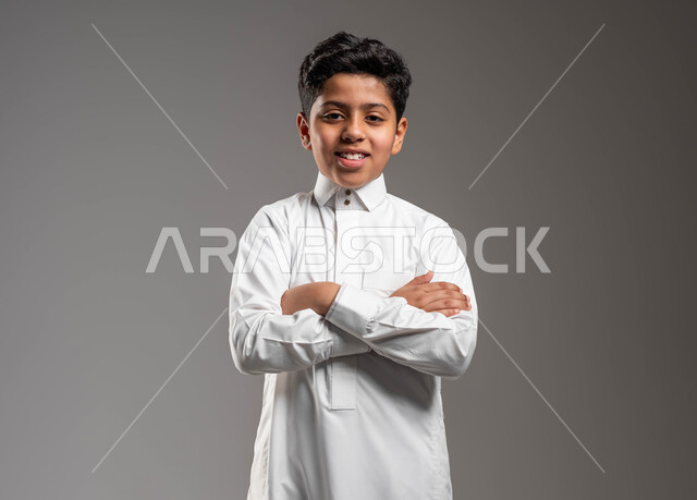 الوقوف باستقامة بإيماءات أيدي متقاطعة، بورتريه لولد عربي خليجي سعودي مبتسم يرتدي الثوب التقليدي ينظر الى الكاميرا بتعابير الفرح والسرور، مفهوم الاهتمام بالمظهر الخارجي, خلفية رمادية