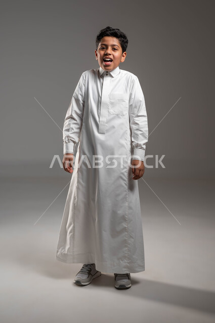 ايماءات الضحك الشديد، بورتريه لولد عربي خليجي سعودي يرتدي الثوب التقليدي وينظر الى الكاميرا بتعابير السخرية والشماتة من شيء ما، صورة الجسم بالطول الكامل، خلفية رمادية