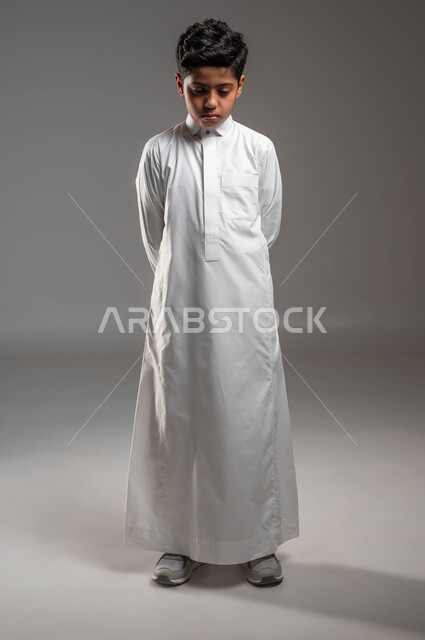 التمعن والتأمل في موضوع ما، ايماءات الحزن والقلق، التفكير في شيء ما، بورتريه لولد عربي خليجي سعودي يرتدي الثوب التقليدي يقف باستقامة وينظر الى الأسفل، ايجاد حل لمشكلة معينة، صورة الجسم بالطول الكامل، خلفية رمادية