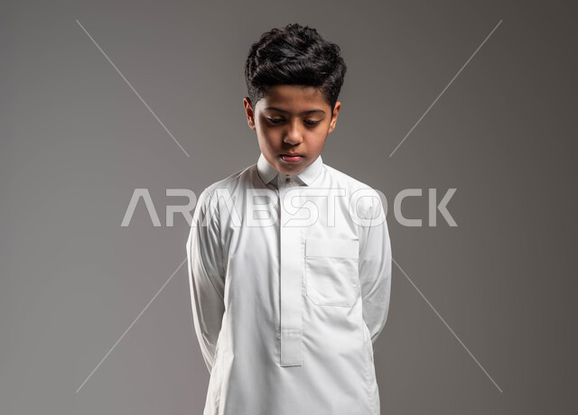 ايماءات الحزن والقلق، التفكير في شيء ما، بورتريه مقرب لولد عربي خليجي سعودي يرتدي الثوب التقليدي يقف باستقامة وينظر الى الأسفل، ايجاد حل لمشكلة معينة، التمعن والتأمل في موضوع ما، خلفية رمادية