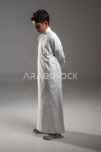 ايجاد حل لمشكلة معيتة، ايماءات الحزن والقلق، التفكير في شيء ما، بورتريه من الجانب لولد عربي خليجي سعودي يرتدي الثوب التقليدي يقف باستقامة وينظر الى الأسفل، التمعن والتأمل في موضوع ما، صورة الجسم بالطول الكامل، خلفية رمادية