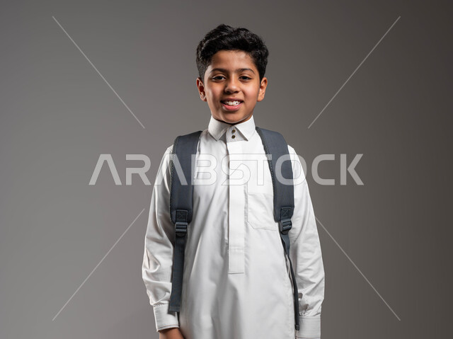 The concept of education in Saudi Arabia and perseverance to achieve success, preparing to go to school, a close-up portrait of a Saudi Gulf Arab student wearing traditional clothing and carrying a school backpack, gestures of joy and happiness for the beginning of the new school year, gray background
