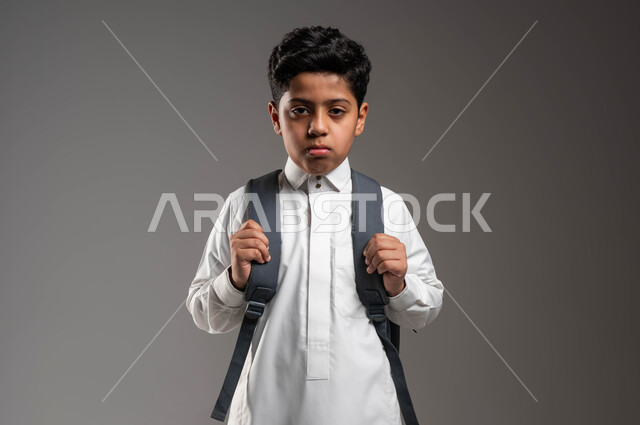 إيماءات الغضب والحزن لبداية العام الدراسي الجديد، عدم الرغبة في الذهاب إلى المدرسة، بورتريه مقرب لتلميذ عربي خليجي سعودي يرتدي الثوب التقليدي يحمل حقيبة الظهر المدرسيه، مفهوم التعليم في المملكة العربية السعودية، خلفية رماديه