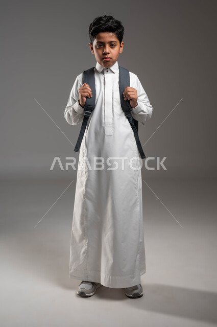 عدم الرغبة في الذهاب إلى المدرسة، بورتريه لتلميذ عربي خليجي سعودي يرتدي الثوب التقليدي يحمل حقيبة الظهر المدرسيه، مفهوم التعليم في المملكة العربية السعودية، إيماءات الغضب والحزن لبداية العام الدراسي الجديد، صورة الجسم بالطول الكامل, خلفية رماديه