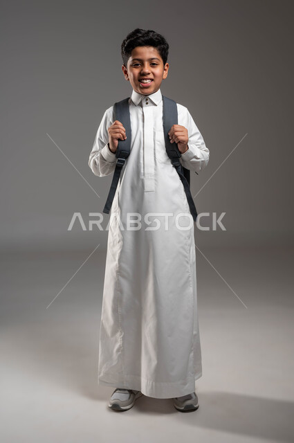 التعليم في السعودية والمثابرة لتحقيق النجاح، الاستعداد للذهاب إلى المدرسة، بورتريه لطالب عربي خليجي سعودي يرتدي الزي التقليدي يحمل حقيبة الظهر المدرسيه، إيماءات الفرح والسعادة لبداية العام الدراسي الجديد، صورة الجسم بالطول الكامل, خلفية رماديه