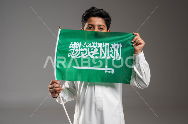 تعليم وتربية الأبناء على حب الوطن والإنتماء إليه, ذكرى اليوم الوطني السعودي ٢٣ سبتمبر، يوم العلم ١١ مارس، بورتريه مقرب لولد عربي خليجي سعودي ينظر إلى الكاميرا يرتدي الزي التقليدي يحمل علم المملكة بفخر وإعتزاز بالهوية الوطنية، خلفية رمادية