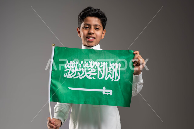 تربية الأبناء على حب الوطن والإنتماء إليه, العيد الوطني السعودي ٢٣ سبتمبر، يوم العلم ١١ مارس، بورتريه مقرب لولد عربي خليجي سعودي مبتسم يرتدي الزي التقليدي  يحمل بيده علم المملكة بفخر وإعتزاز بالهوية الوطنية، النظر إلى الكاميرا بإيماءات السرور، خلفية رمادية