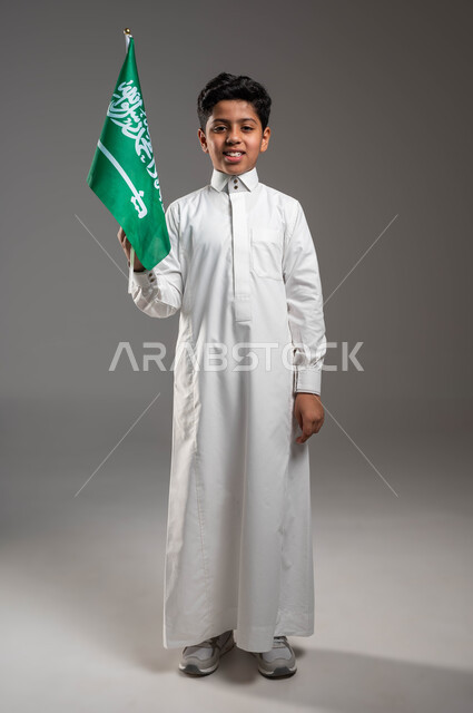 النظر إلى الكاميرا بإيماءات السرور، يوم العلم ١١ مارس، بورتريه لولد عربي خليجي سعودي مبتسم يرتدي الزي التقليدي يحمل علم المملكة بفخر وإعتزاز بالهوية الوطنية، ذكرى اليوم الوطني السعودي ٢٣ سبتمبر، صورة الجسم بالطول الكامل, خلفية رمادية