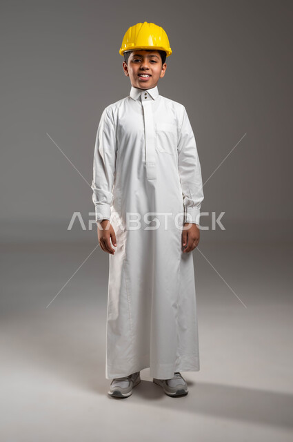 الشغف بمهنة الهندسة منذ الطفولة، مفهوم الهندسة المدنية والمعمارية، بورتريه لفتى عربي خليجي سعودي مبتسم يرتدي الثوب التقليدي و الخوذة الوقاية الخاصة بالعمل ينظر إلى الكاميرا، مهن ووظائف مستقبلية، صورة الجسم بالطول الكامل، خلفية رمادية