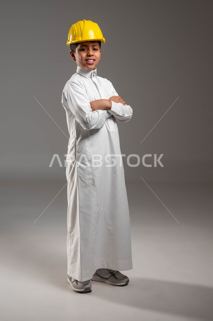 مهن ووظائف مستقبلية، الشغف بمهنة الهندسة منذ الطفولة، بورتريه لولد عربي خليجي سعودي يرتدي الثوب التقليدي وخوذة الحماية الخاصة بالعمل، يقف بأيدي متقاطعة، تعابير السرور لتحقيق حلم النجاح، صورة الجسم بالطول الكامل، خلفية رمادية