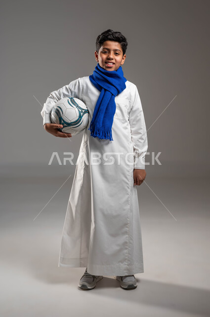 جاهز للتحدي والفوز، بورتريه لولد عربي خليجي سعودي يرتدي الثوب التقليدي ووشاح أزرق يحمل بيده كرة قدم بتعابير الفرح والسرور والحماس للمنافسة، تشجيع الفريق السعودي، خلفية رمادية، النظر إلى الكاميرا بروح العزيمة على الانتصار، صورة الجسم بالطول الكامل