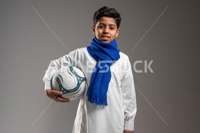 جاهز للتحدي والفوز، بورتريه مقرّب لولد عربي خليجي سعودي يرتدي الثوب التقليدي ووشاح أزرق يحمل بيده كرة قدم بتعابير الفرح والسرور والحماس للمنافسة، تشجيع الفريق السعودي، خلفية رمادية، النظر إلى الكاميرا بروح العزيمة على الانتصار