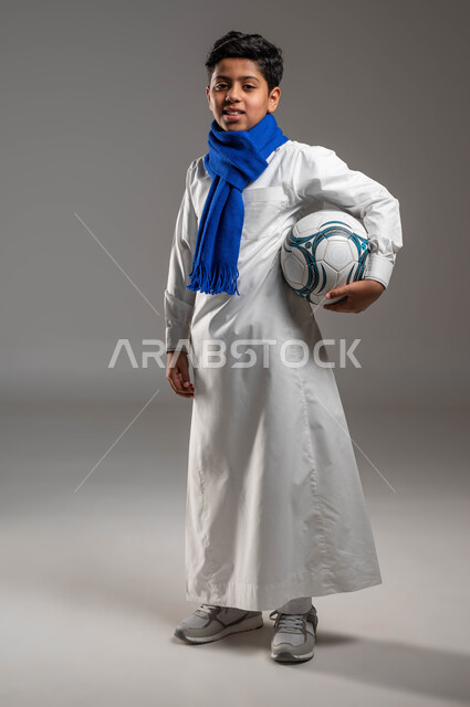 جاهز للتحدي والفوز، بورتريه لولد عربي خليجي سعودي يرتدي الثوب التقليدي ووشاح أزرق يحمل بيده كرة قدم بتعابير الفرح والسرور والحماس للمنافسة، تشجيع الفريق السعودي، خلفية رمادية، النظر إلى الكاميرا بروح العزيمة على الانتصار، صورة الجسم بالطول الكامل