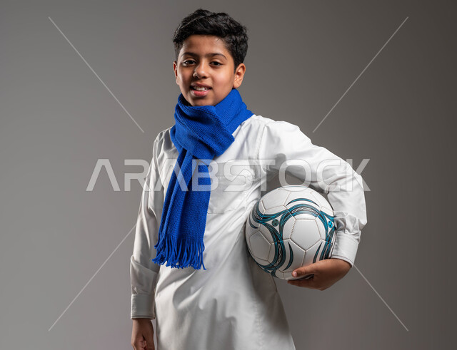 جاهز للتحدي والفوز، بورتريه مقرّب لولد عربي خليجي سعودي يرتدي الثوب التقليدي ووشاح أزرق يحمل بيده كرة قدم بتعابير الفرح والسرور والحماس للمنافسة، تشجيع الفريق السعودي، خلفية رمادية، النظر إلى الكاميرا بروح العزيمة على الانتصار