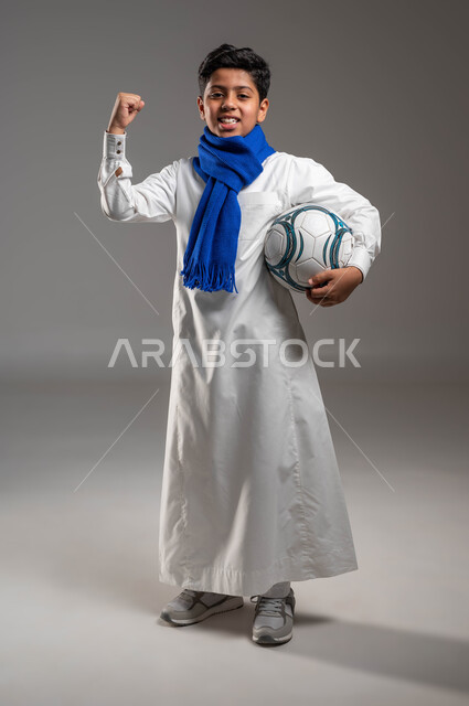 إيماءات الفوز والانتصار والقوة، بورتريه لولد عربي خليجي سعودي يرتدي الثوب التقليدي ووشاح أزرق يرفع يده للاعلى بتعابير الفرح والسرور ويحمل بيده كرة قدم، تشجيع الفريق السعودي، خلفية رماديه، النظر إلى الكاميرا ، صورة الجسم بالطول الكامل