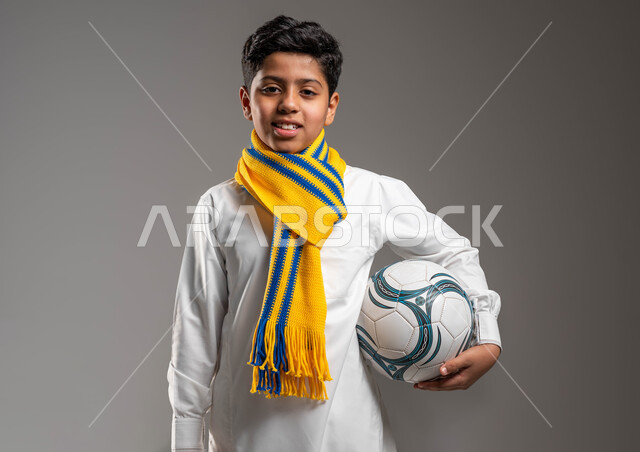 جاهز للتحدي والفوز، بورتريه مقرّب لولد عربي خليجي سعودي يرتدي الثوب التقليدي ووشاح أصفر يحمل بيده كرة قدم بتعابير الفرح والسرور والحماس للمنافسة، تشجيع الفريق السعودي، خلفية رمادية، النظر إلى الكاميرا بروح العزيمة على الانتصار