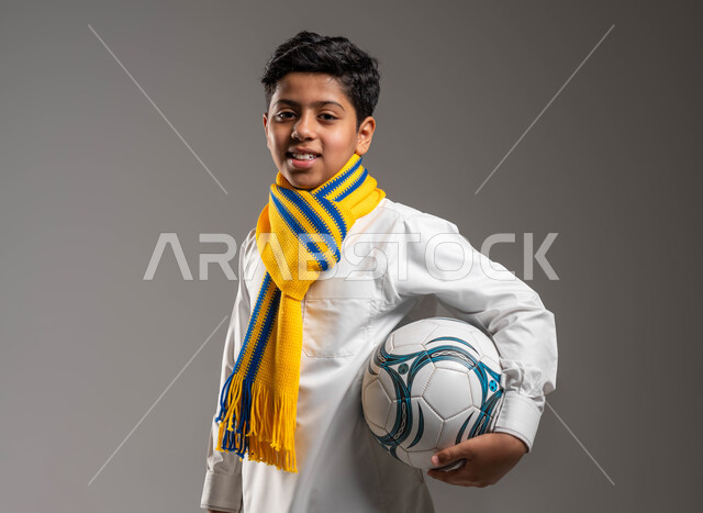 يرتدي الثوب التقليدي ووشاح أصفر يحمل بيده كرة قدم بتعابير الفرح والسرور والحماس للمنافسة، بورتريه مقرّب لولد عربي خليجي سعودي ،تشجيع الفريق السعودي، النظر إلى الكاميرا بروح العزيمة على الانتصار، جاهز للتحدي والفوز، خلفية رمادية