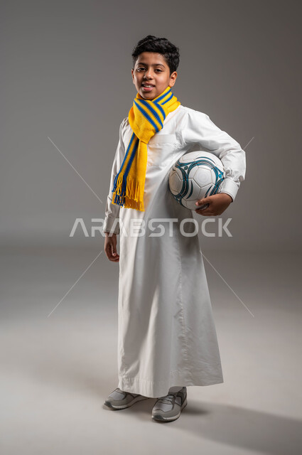 النظر إلى الكاميرا بروح العزيمة على الانتصار، صورة الجسم بالطول الكامل، جاهز للتحدي والفوز، بورتريه لولد عربي خليجي سعودي يرتدي الثوب التقليدي ووشاح أصفر يحمل بيده كرة قدم بتعابير الفرح والسرور والحماس للمنافسة، تشجيع الفريق السعودي، خلفية رمادية