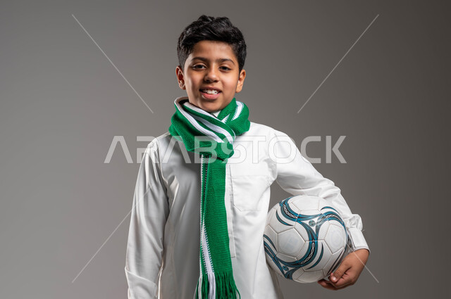 Passion for following sports clubs, a close-up portrait of a smiling Saudi Gulf Arab boy wearing a traditional dress, holding a football in his hand, wearing scarves to encourage the favorite team, looking at the camera with gestures of determination to achieve victory and triumph, gray background.